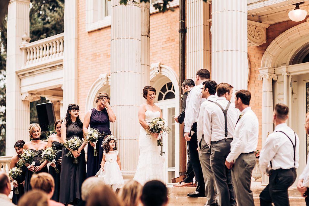 Wedding party assembles on grand stone steps of historic mansion during outdoor ceremony.