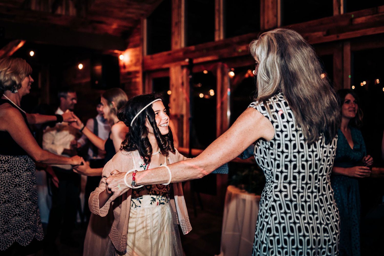 Wedding guests dance together at a rustic indoor reception with wooden beams and warm lighting.