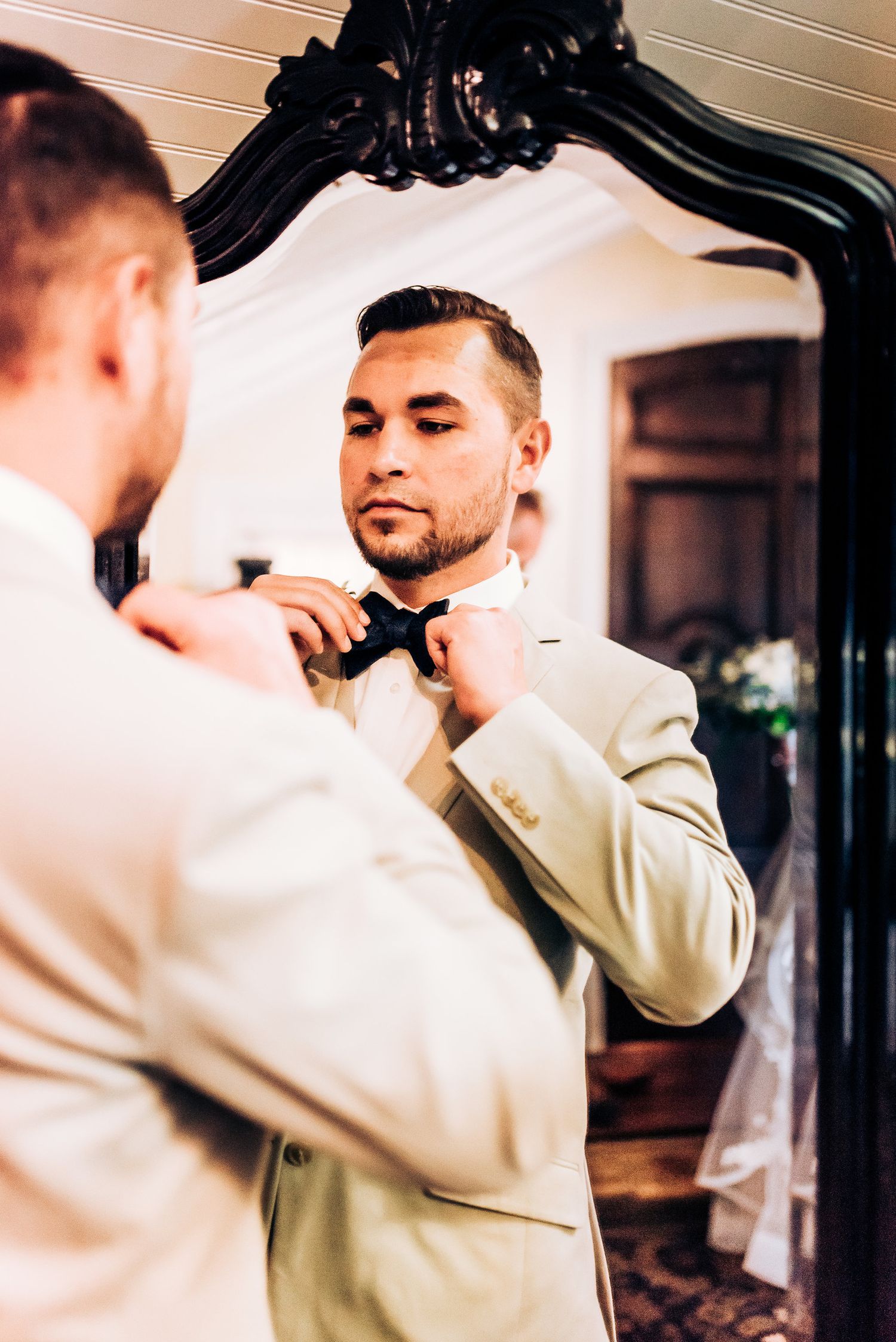 Groom adjusting bow tie while looking in ornate mirror before ceremony.
