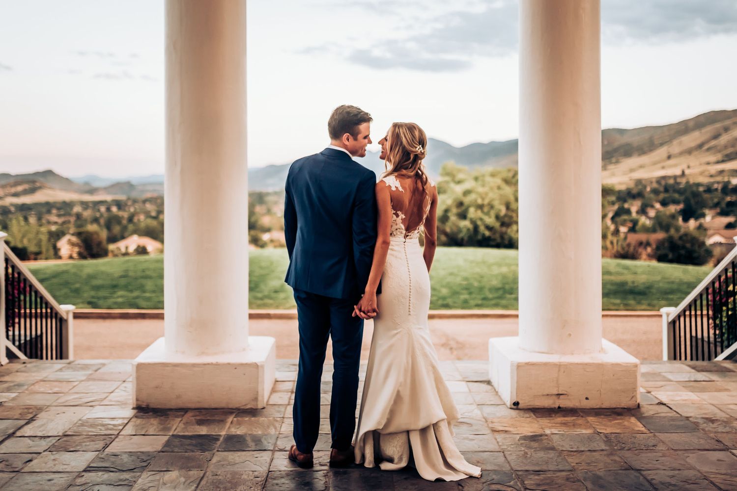 A couple in wedding attire stands between pillars overlooking a mountain landscape.