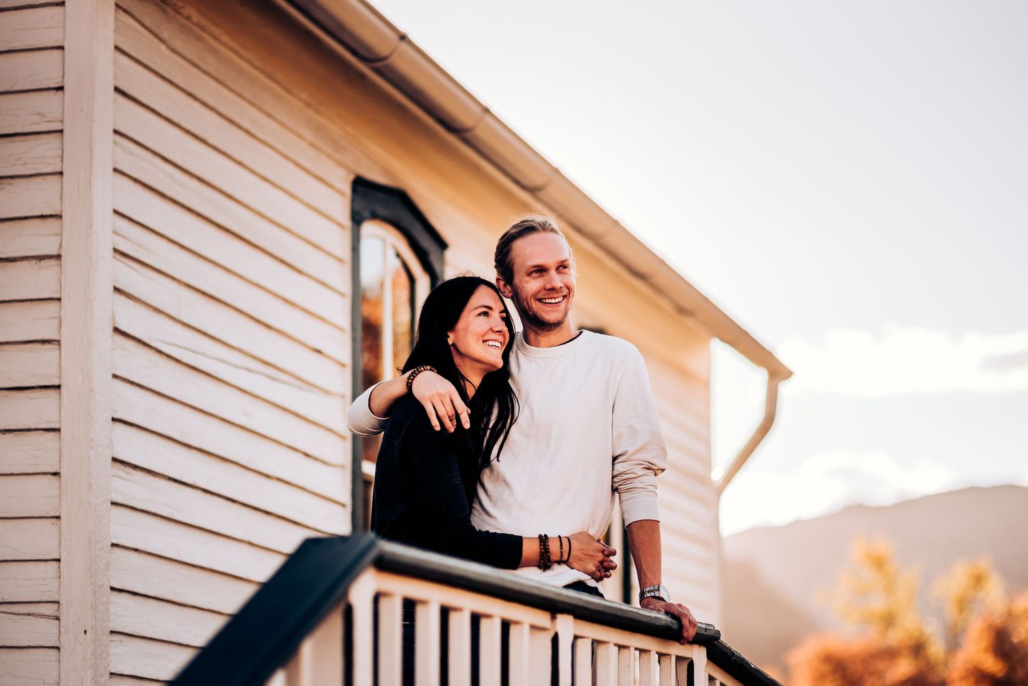 A couple stands together on a porch railing with mountains and sunset in the background.