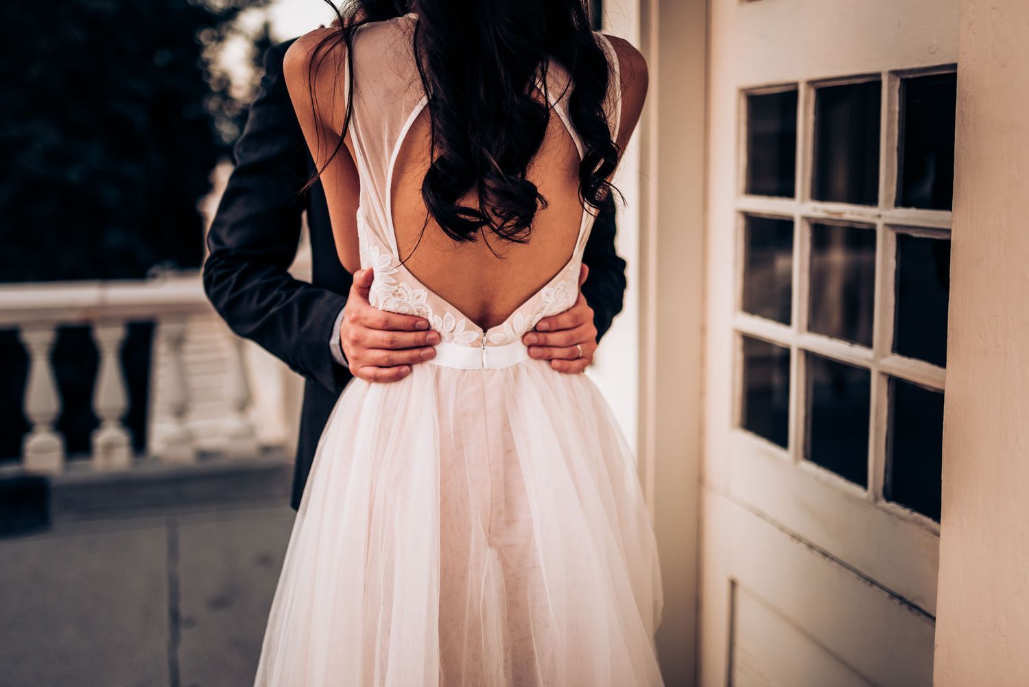A romantic wedding photo showing the back of a white tulle dress with a groom's hands on the bride's waist.