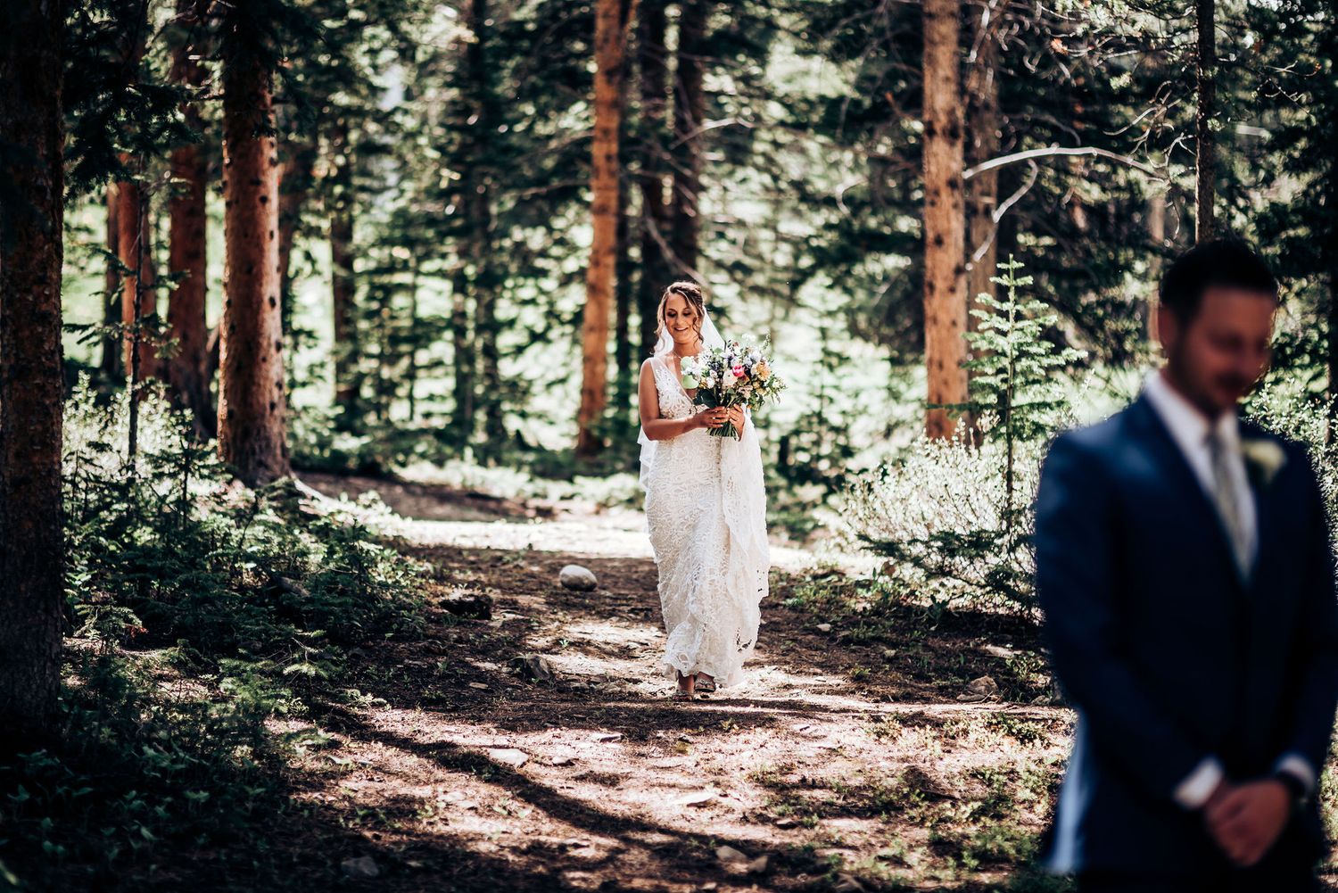 A bride in white stands between tall pine trees as sunlight filters through the forest canopy.