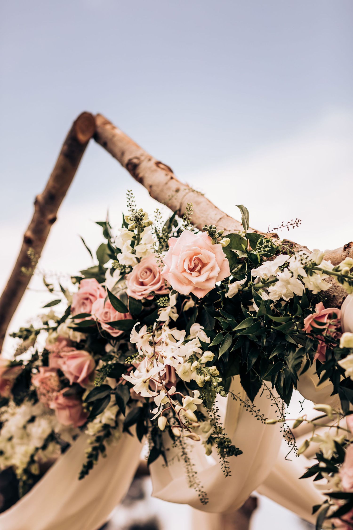 Rustic wooden arch decorated with blush roses and white flowers against cloudy sky.