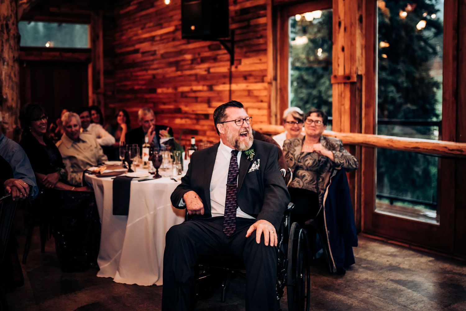 Wedding guest in formal attire sits at a rustic wooden venue with tables and windows at dusk.