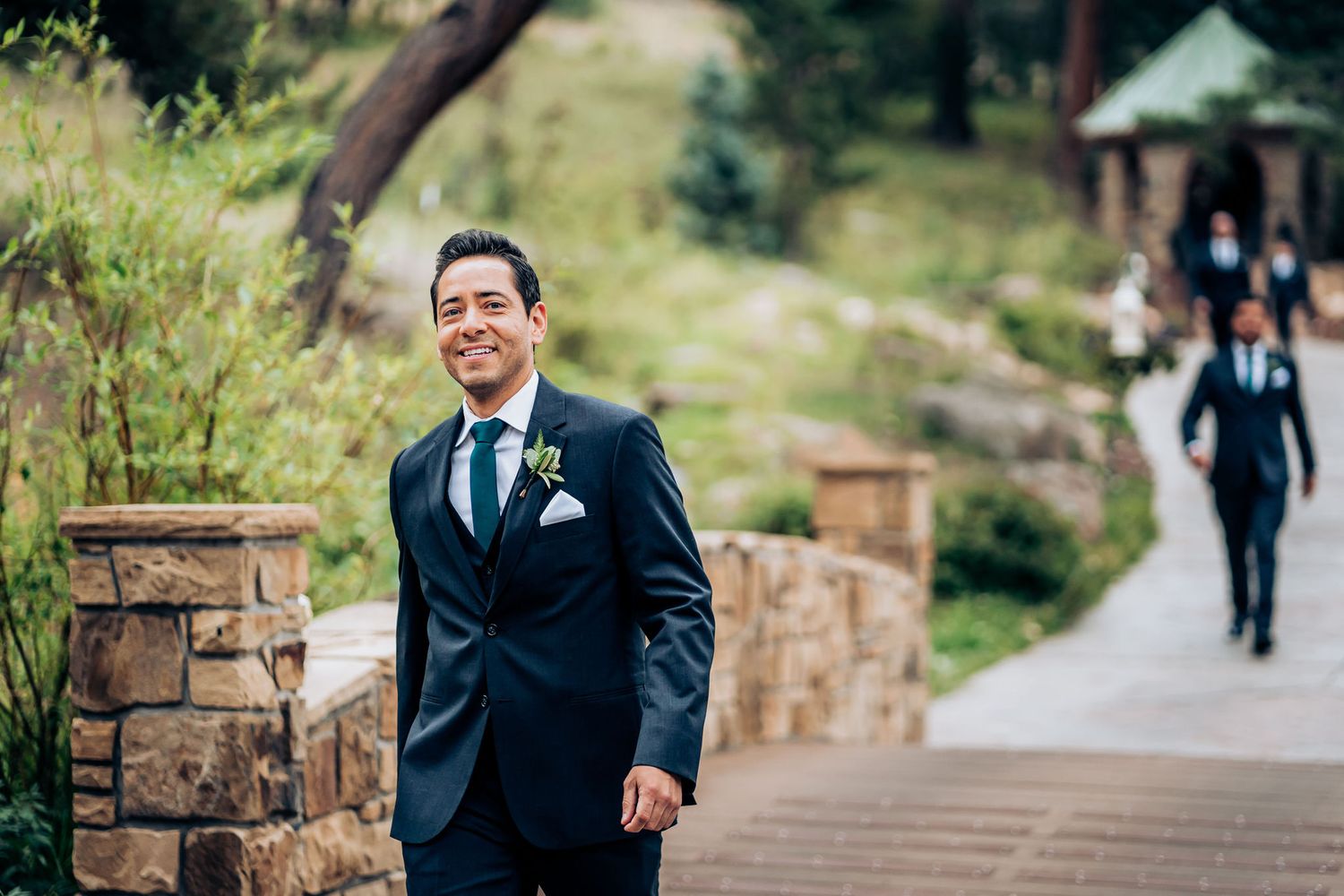 Person in navy suit smiling while standing by stone wall on outdoor path.