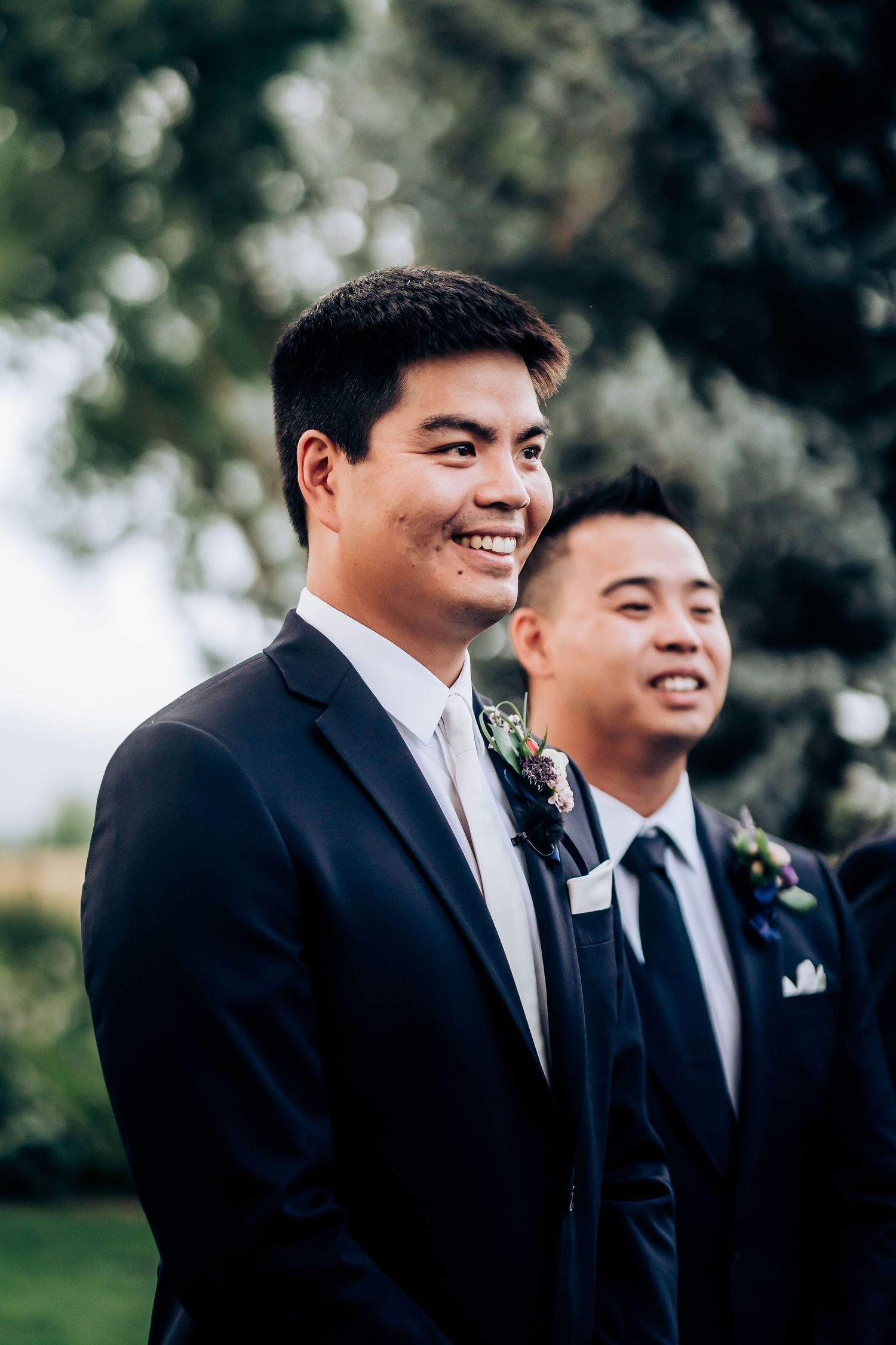 Groomsmen in dark suits with boutonnieres smile during outdoor wedding ceremony.