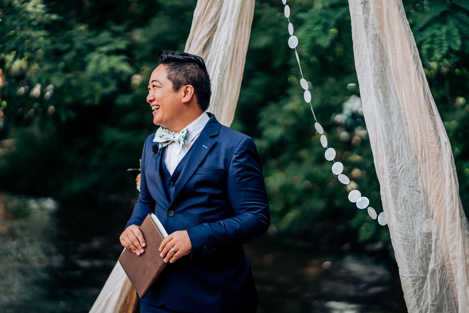 Groom in a navy blue suit stands thoughtfully near draped curtains at an outdoor ceremony.