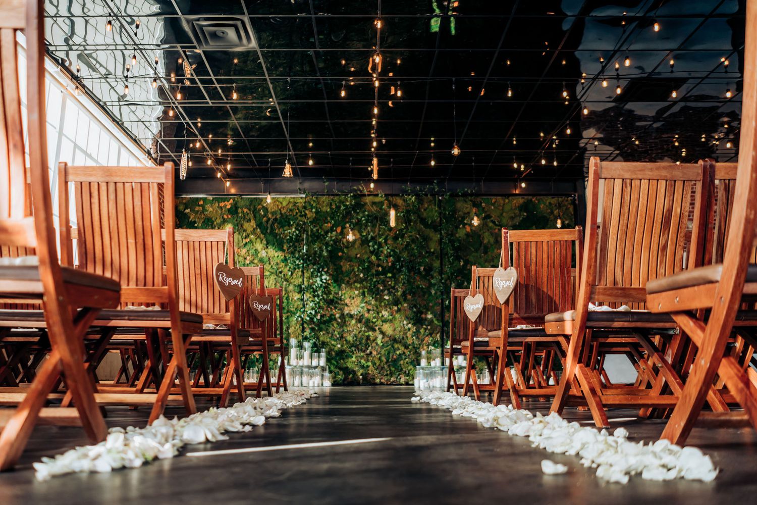 Wedding ceremony space with wooden chairs and greenery-lined aisle under string lights.