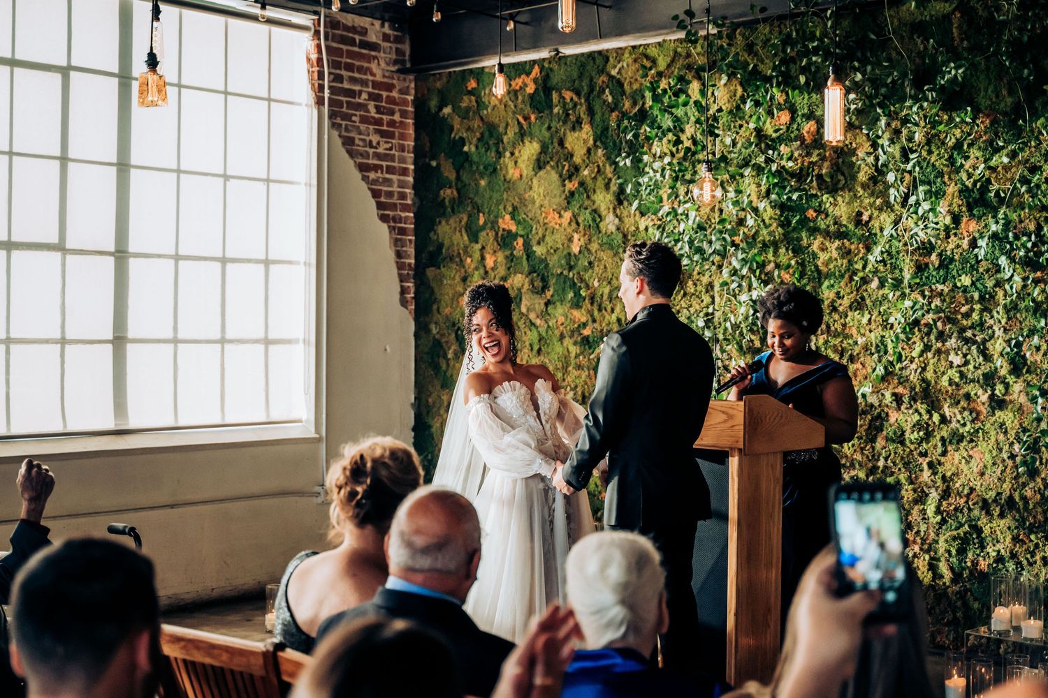 Indoor wedding ceremony with guests watching couple exchange vows against foliage wall.
