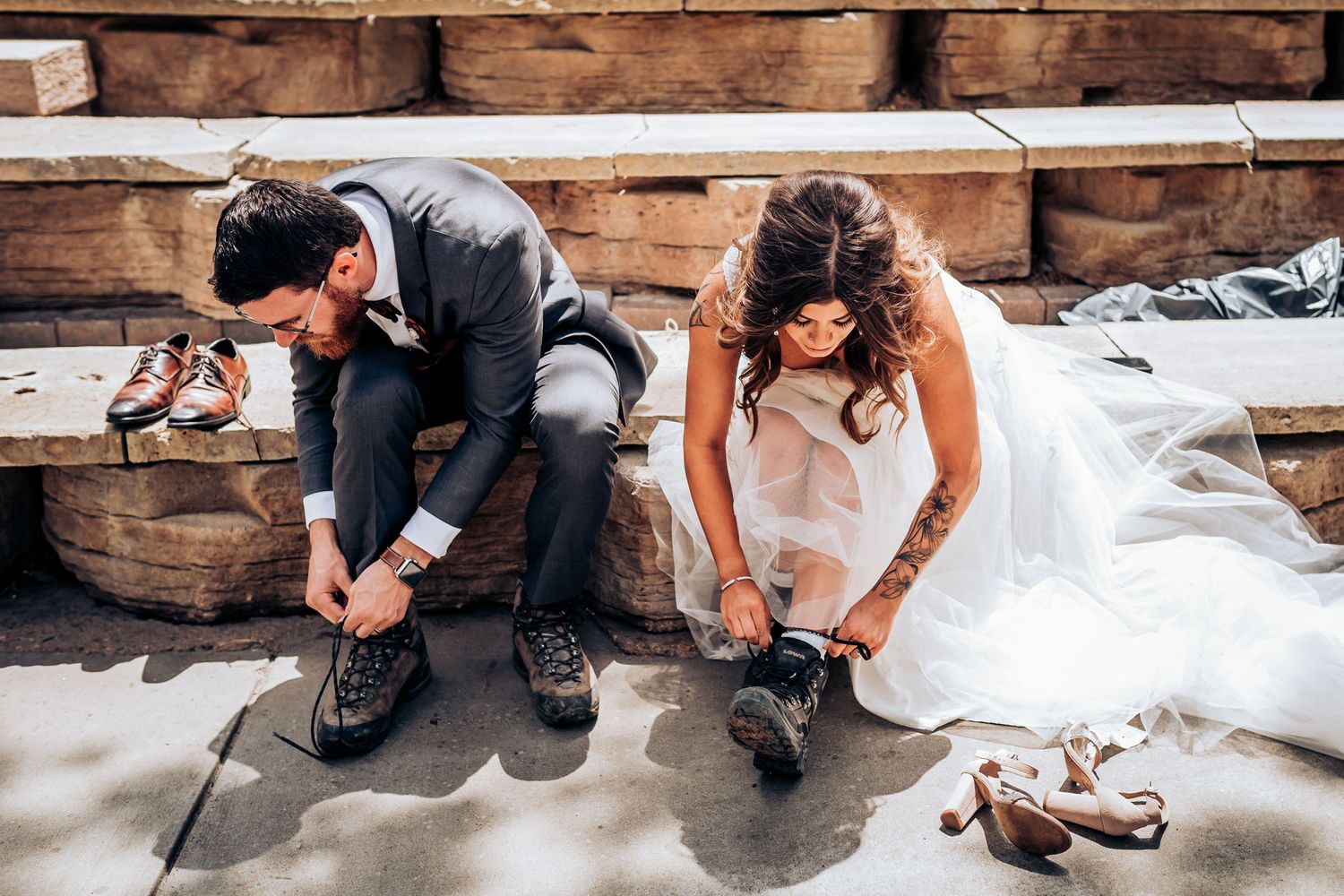 Wedding couple changing into hiking boots while sitting on wooden steps outdoors.