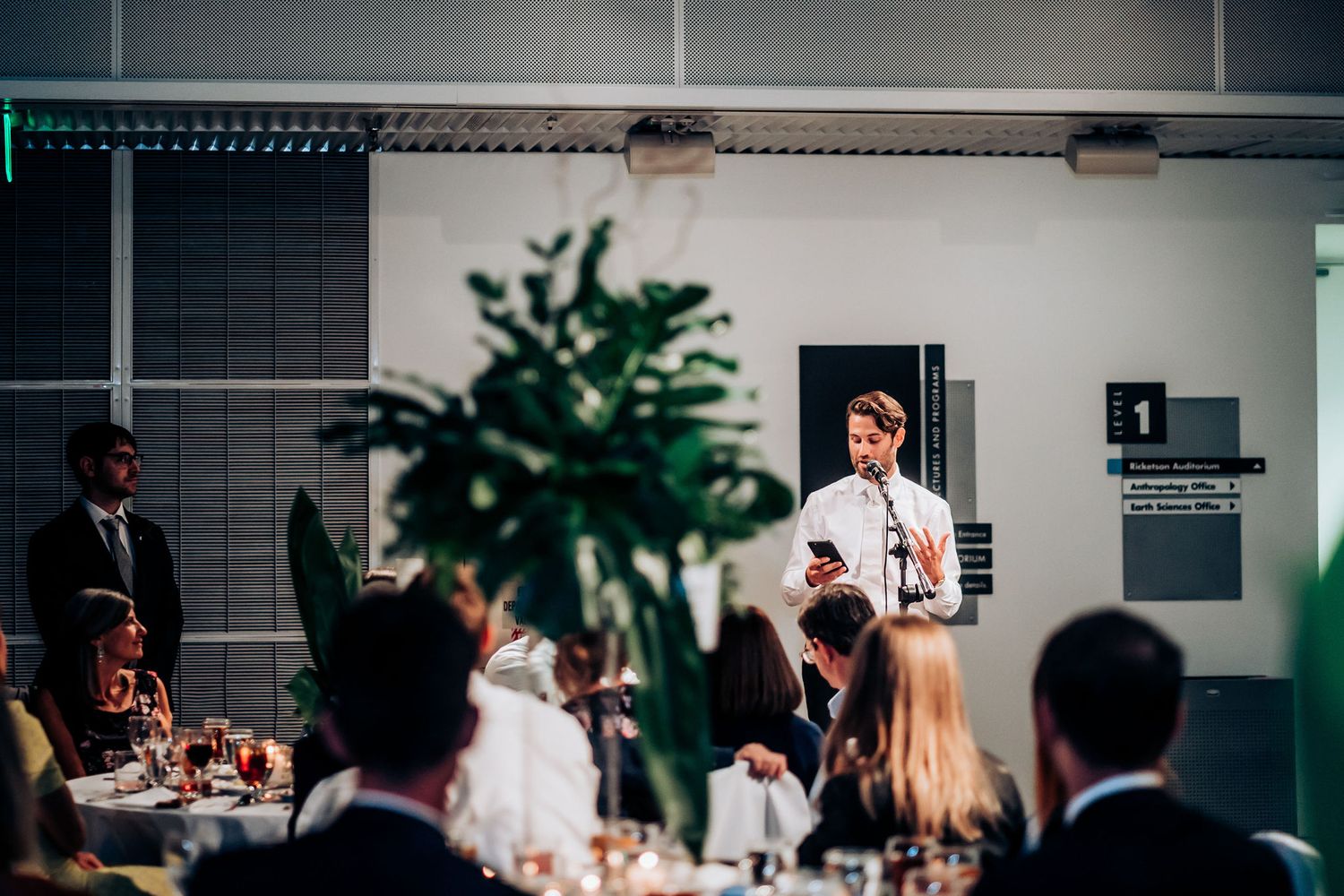 A speaker addresses wedding guests during a reception with decorative plants in the foreground.