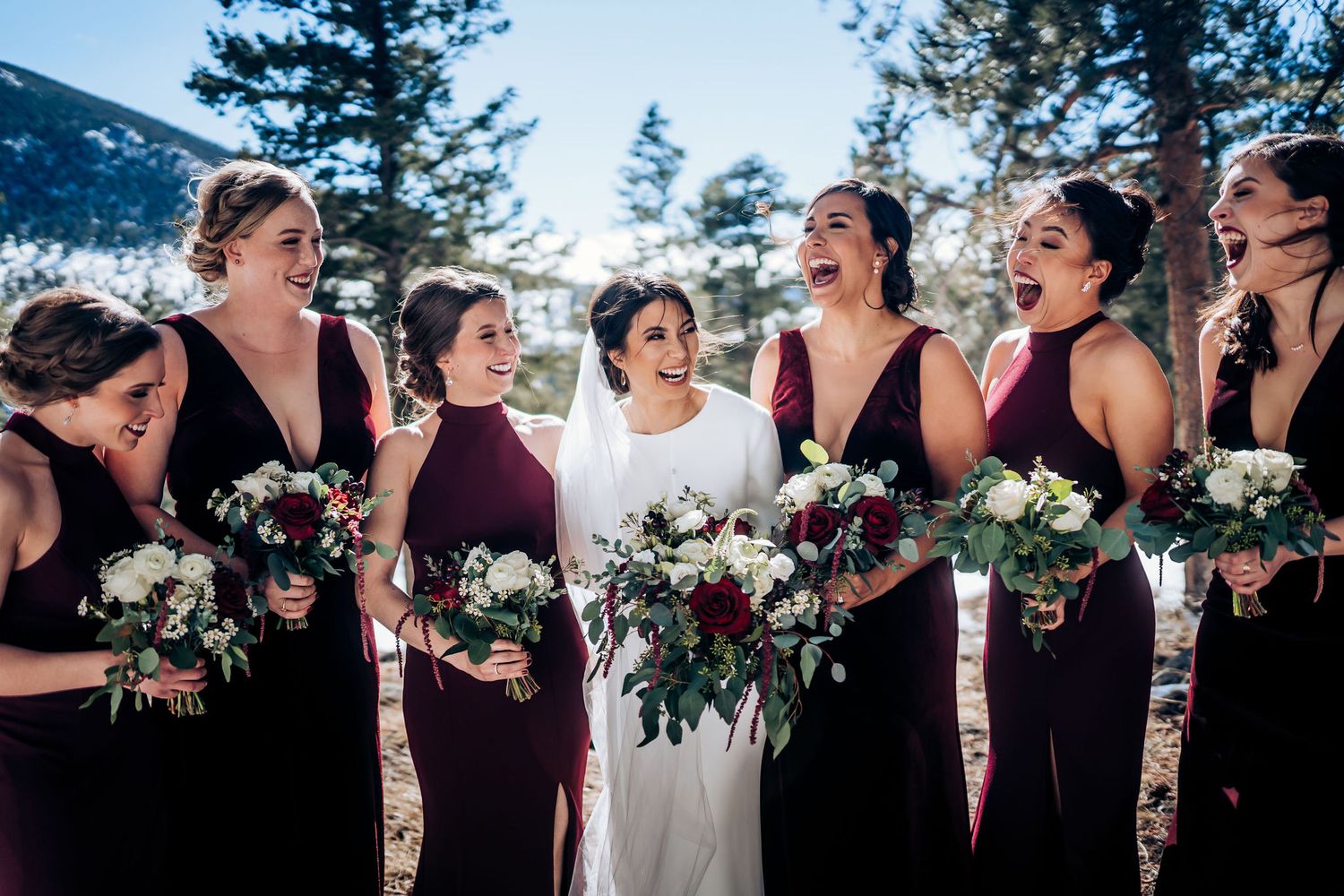 Wedding party laughs together in burgundy dresses holding winter floral bouquets in mountain setting.