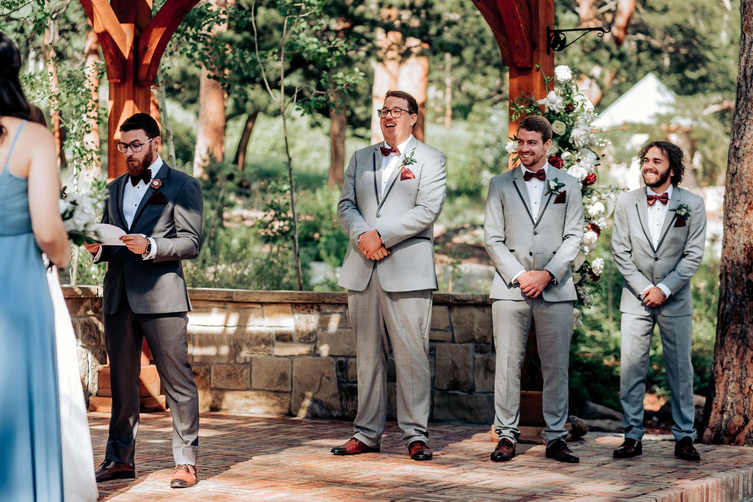 Groomsmen in gray suits stand under a wooden pavilion during an outdoor ceremony.