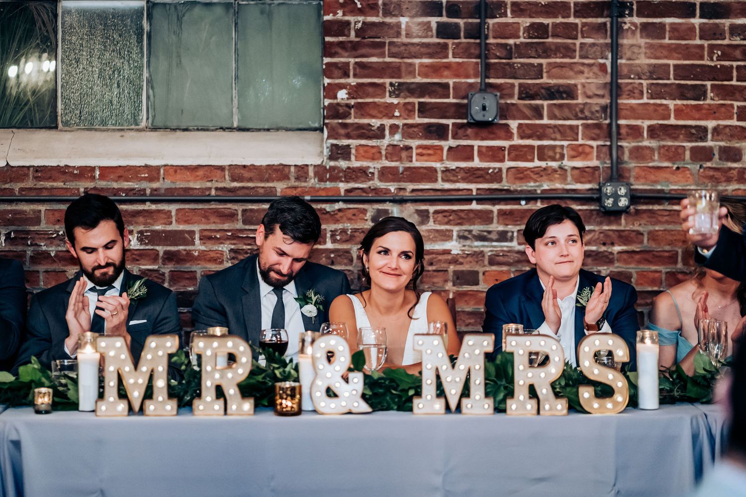 Wedding party seated at head table with illuminated 'MR & MRS' marquee letters as decor.