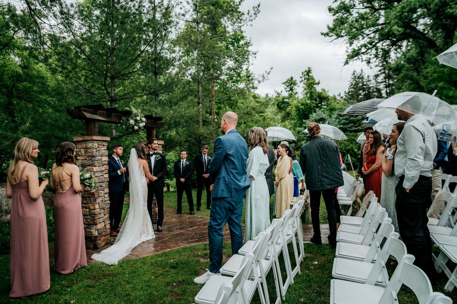 Wedding ceremony takes place outdoors with white chairs and stone pillars surrounded by lush greenery.