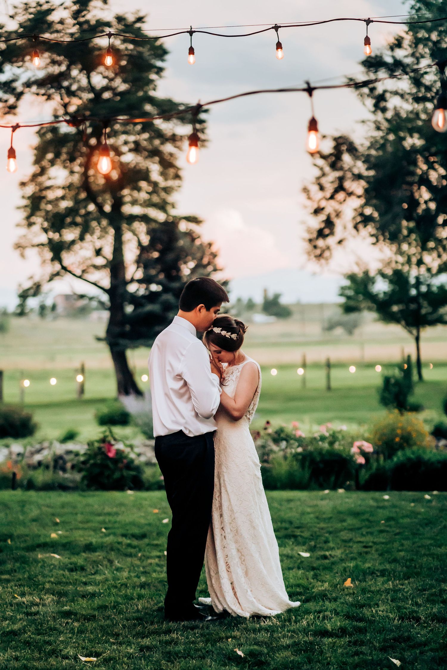 A romantic moment under string lights at dusk in a garden setting with trees and flowers.