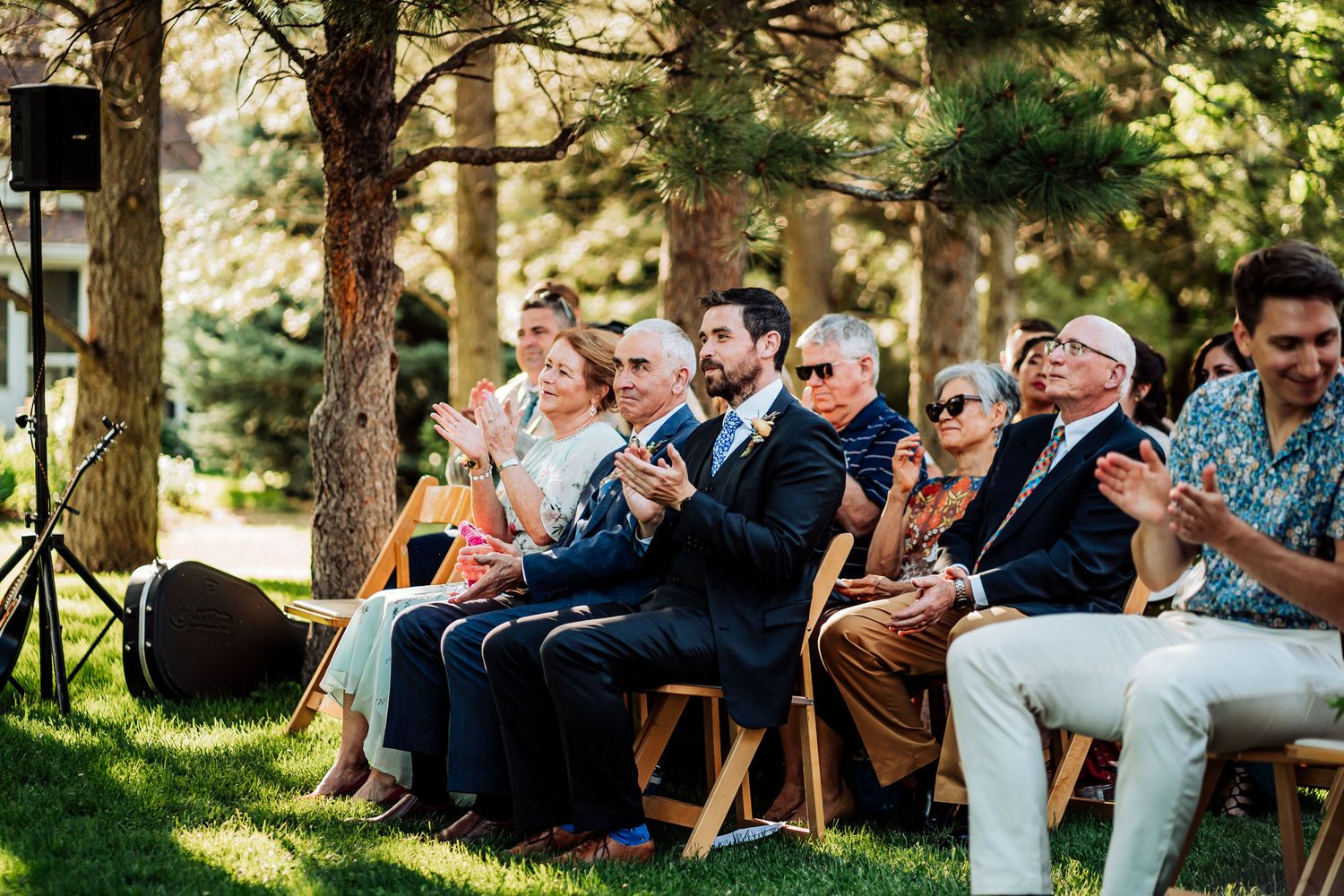 Wedding guests seated outdoors at a garden ceremony smile and watch the proceedings on a sunny day.