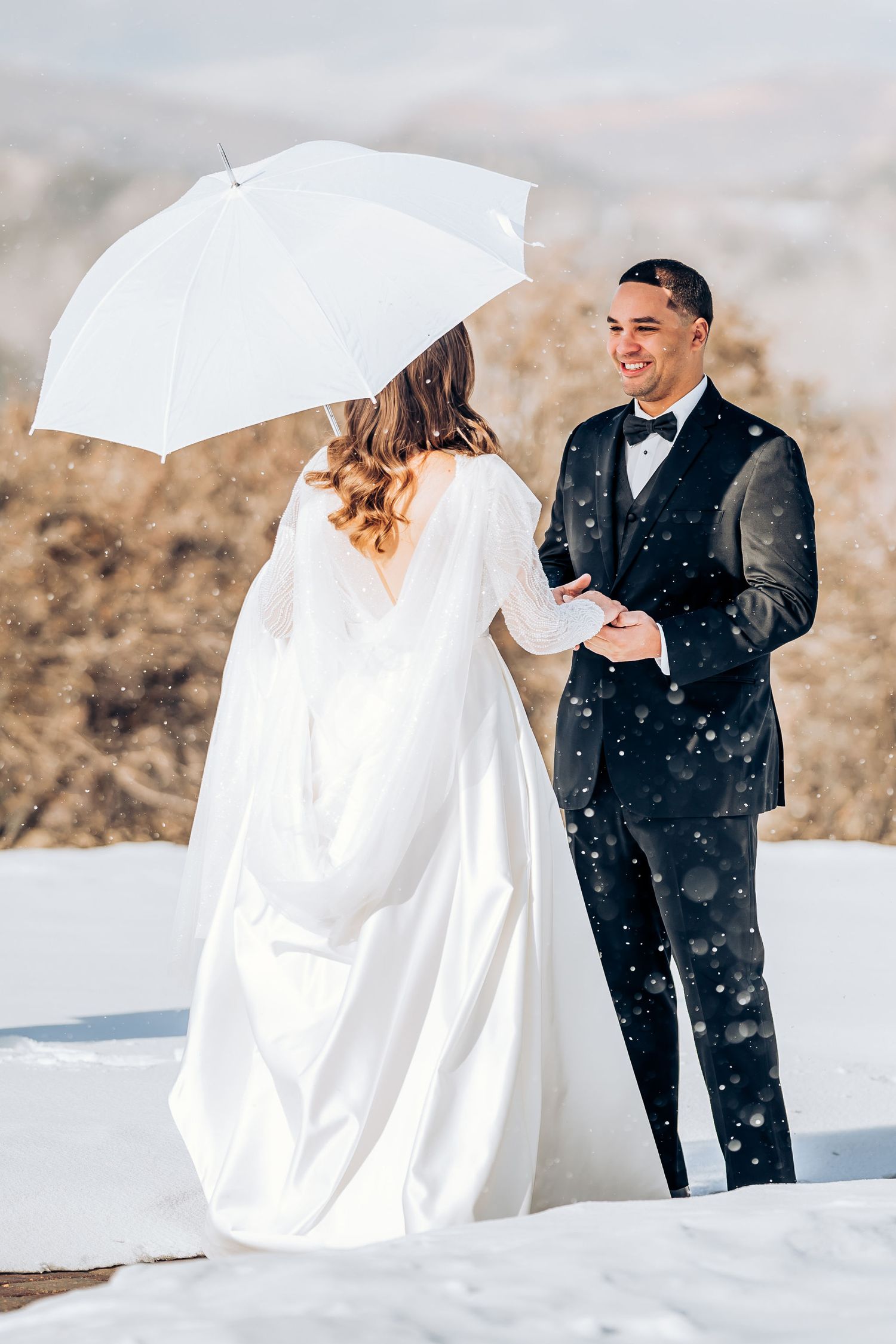 A joyful winter wedding moment captured with falling snow and a white umbrella.