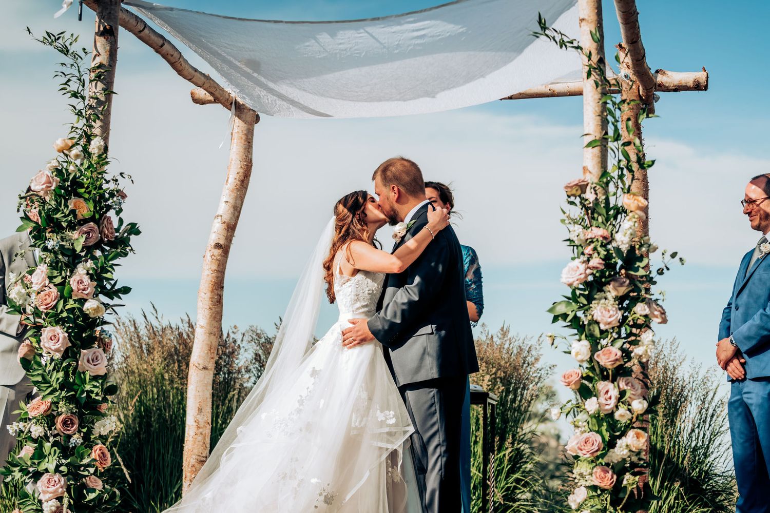 A romantic wedding ceremony under a rustic floral arch with white fabric draped overhead in an outdoor setting.