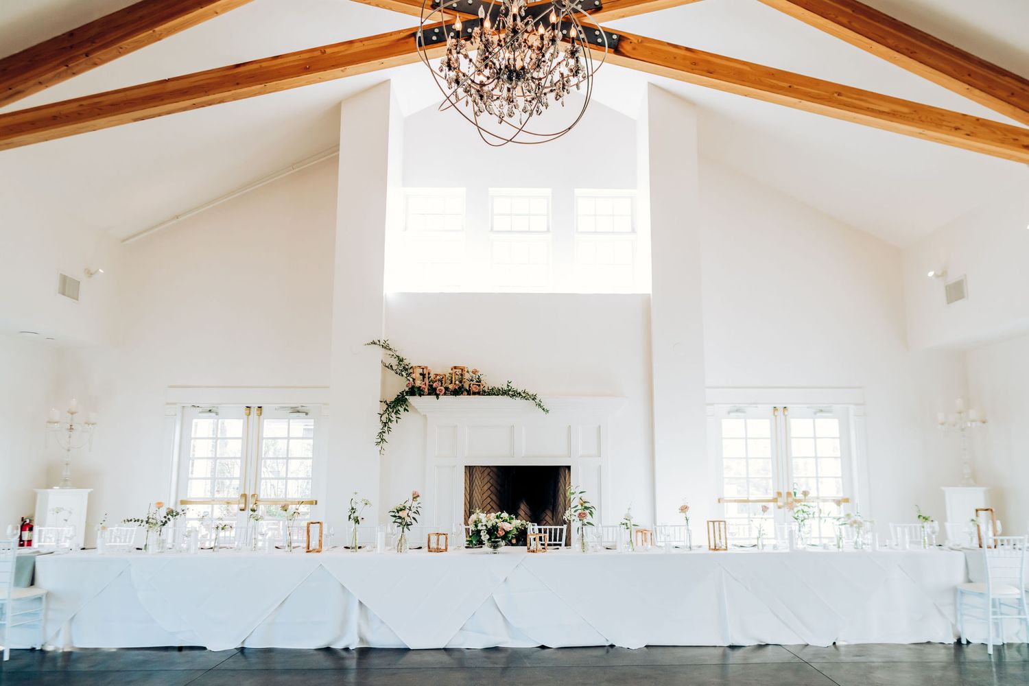 Elegant white wedding reception venue with vaulted ceiling, chandelier, and head table decorated in white linens.