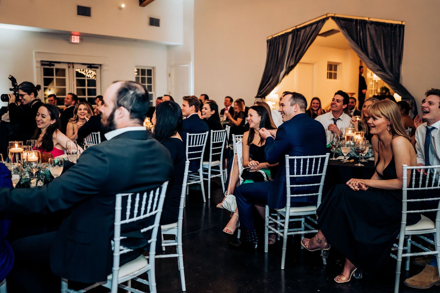 Wedding guests seated at reception tables in elegant venue with high ceilings and chandeliers.