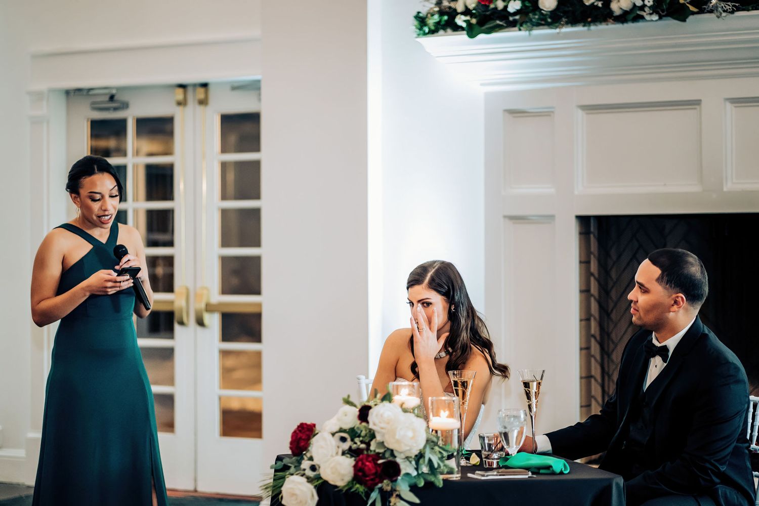 Guest delivers wedding speech at head table during elegant indoor reception with white decor.