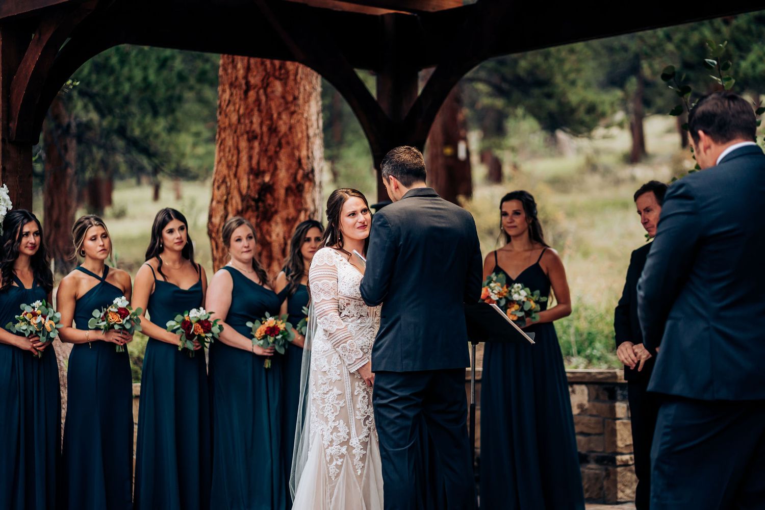 Wedding ceremony framed by wooden pergola with bridal party in navy blue and formal attire.