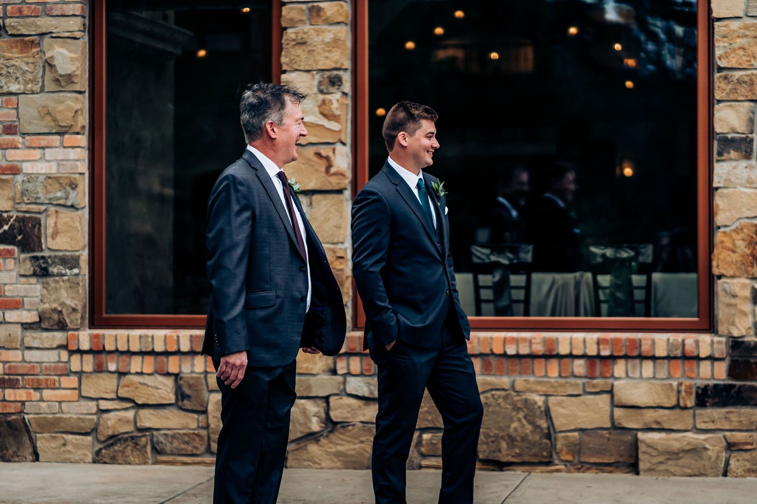 Two men in dark suits walking outside stone building during wedding event.