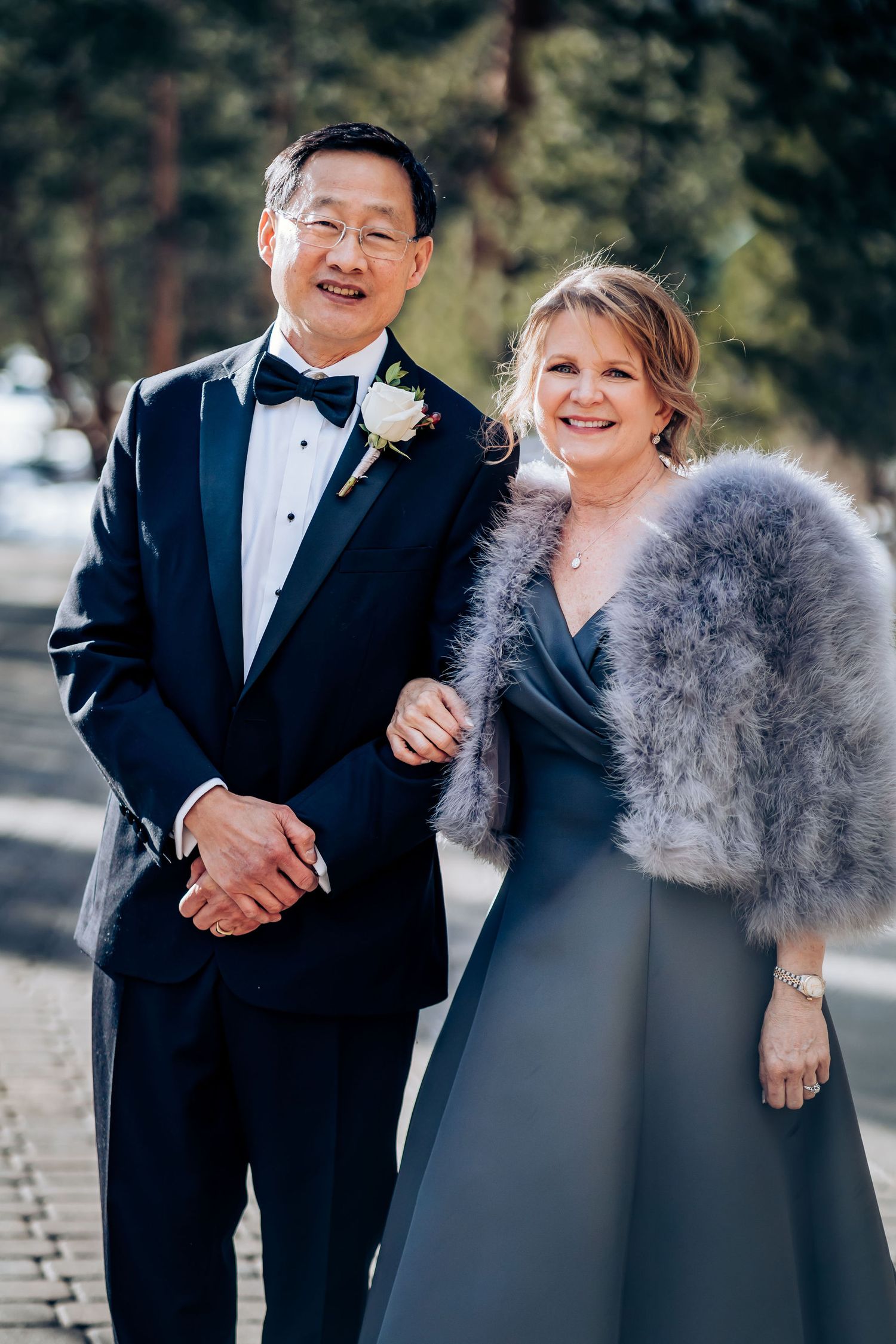 Elegant couple in formal attire at an outdoor wedding event, wearing a tuxedo and grey gown with fur wrap.
