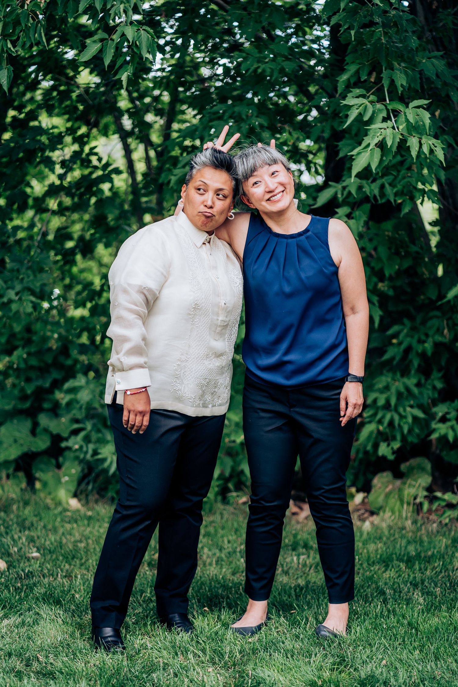 Two people in contrasting outfits share a playful moment on grass with lush greenery behind them.