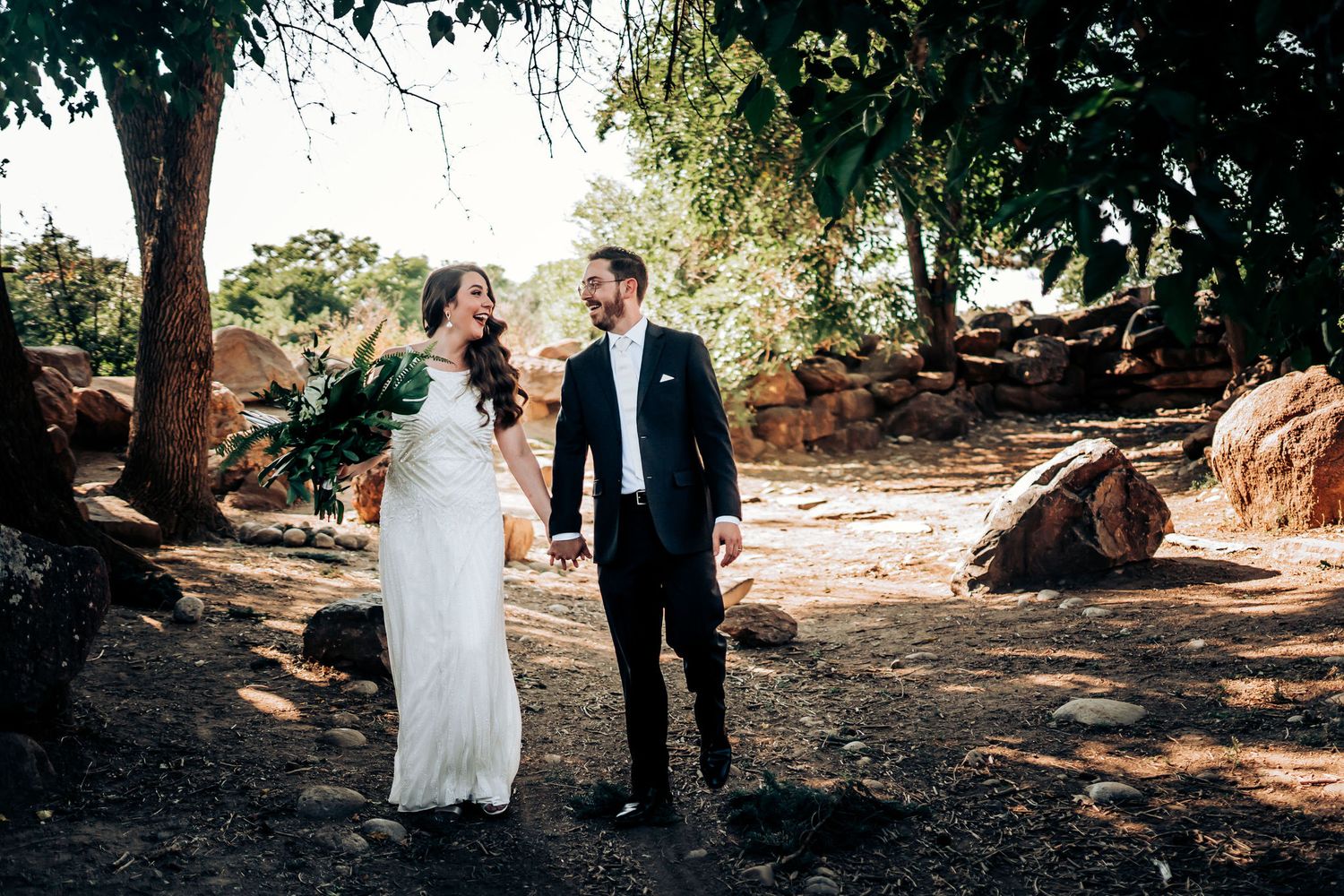 A romantic wedding couple walking together on a tree-lined path in formal attire.