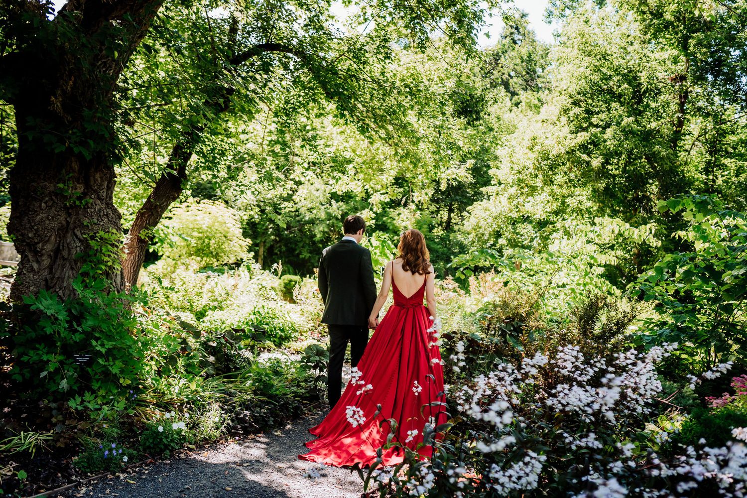 A couple walking along a garden path, with a flowing red dress trailing behind.