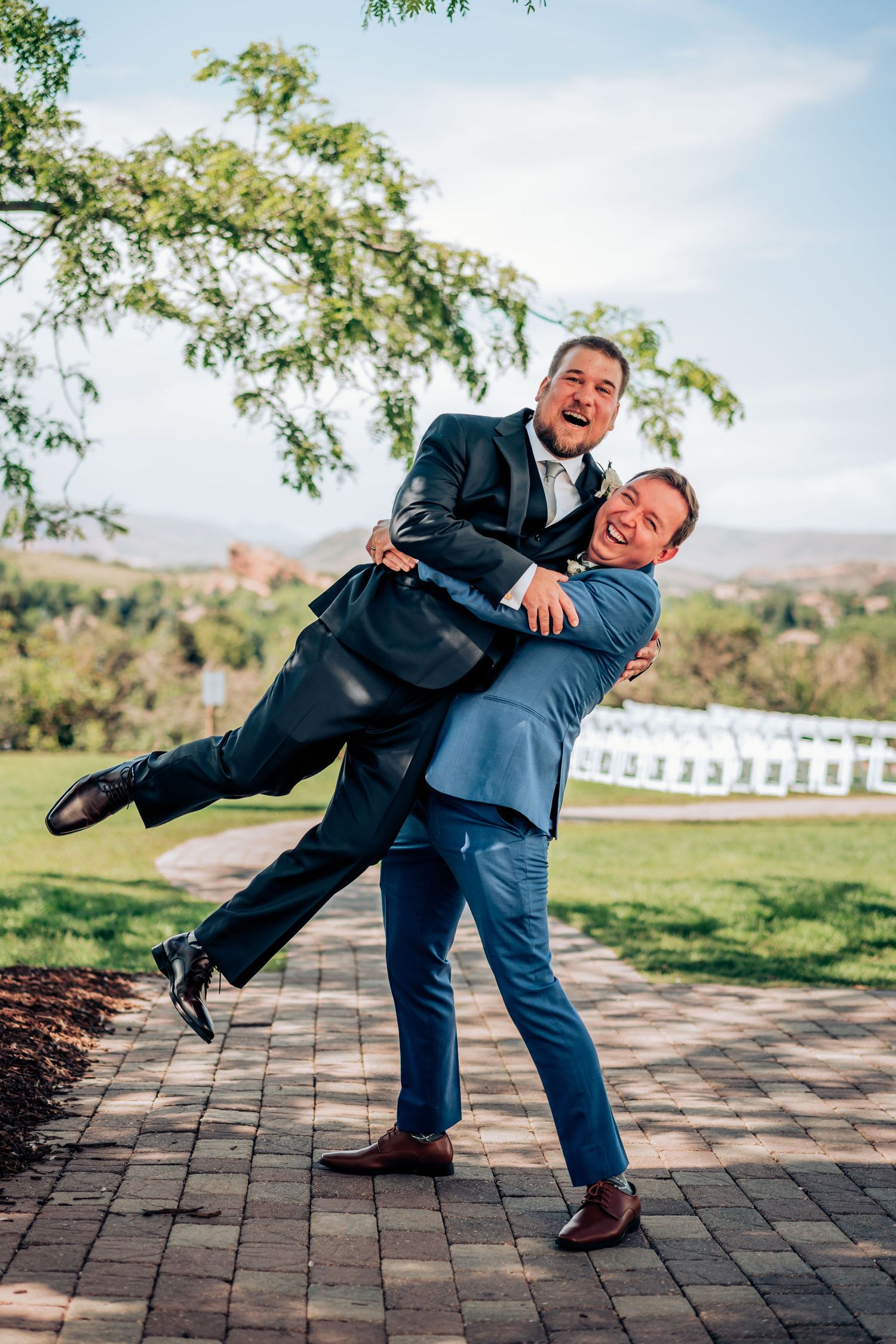Playful groomsmen in suits horsing around on outdoor brick patio at wedding venue.