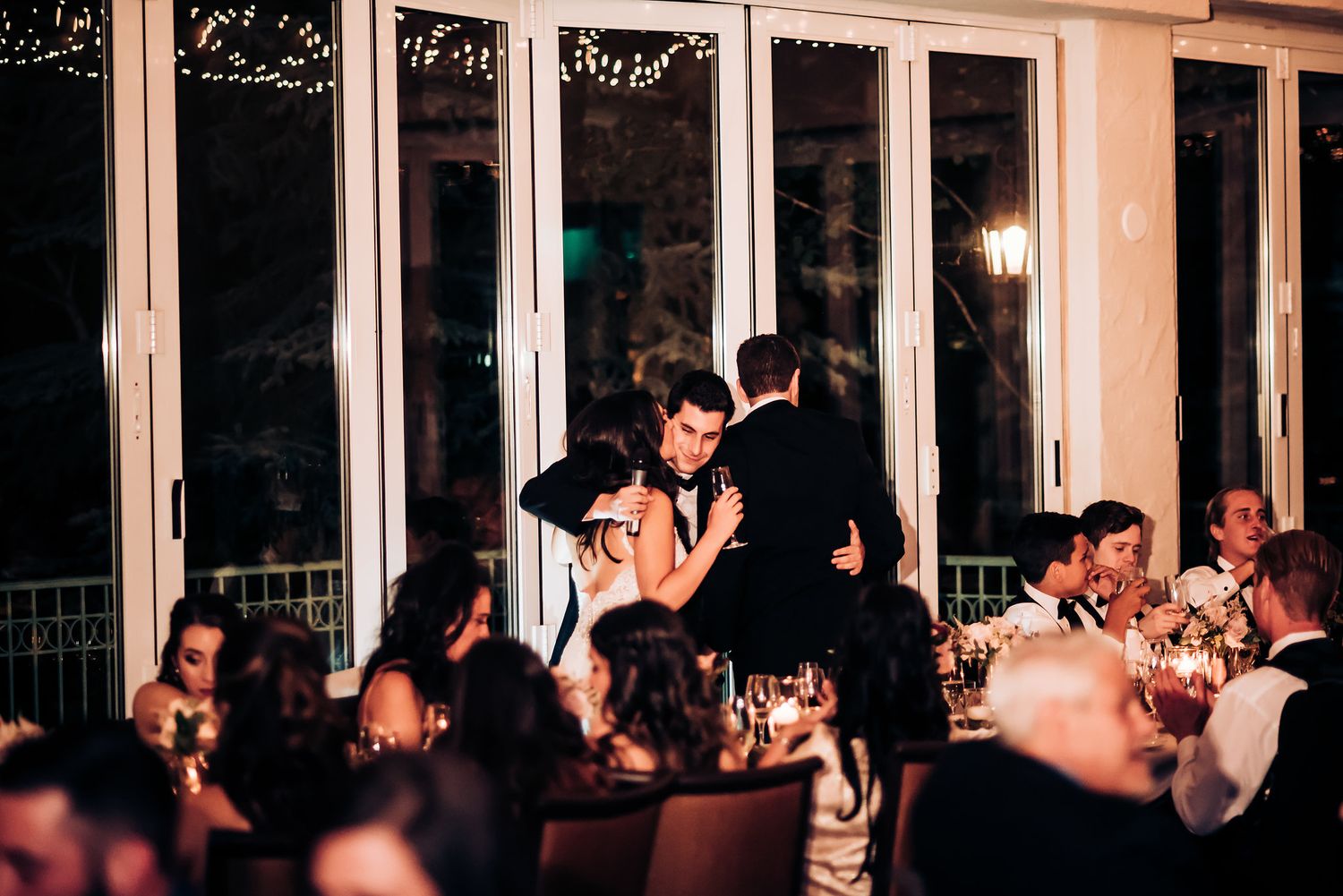 Wedding guests celebrate during an evening reception in a venue with dramatic floor-to-ceiling windows.