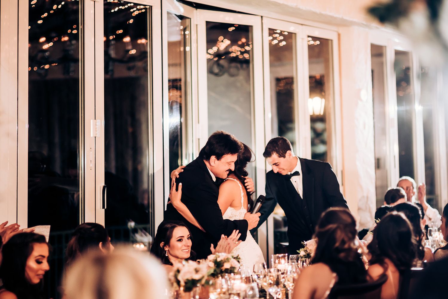 Elegant wedding reception scene with guests mingling at candlelit tables near glass windows.