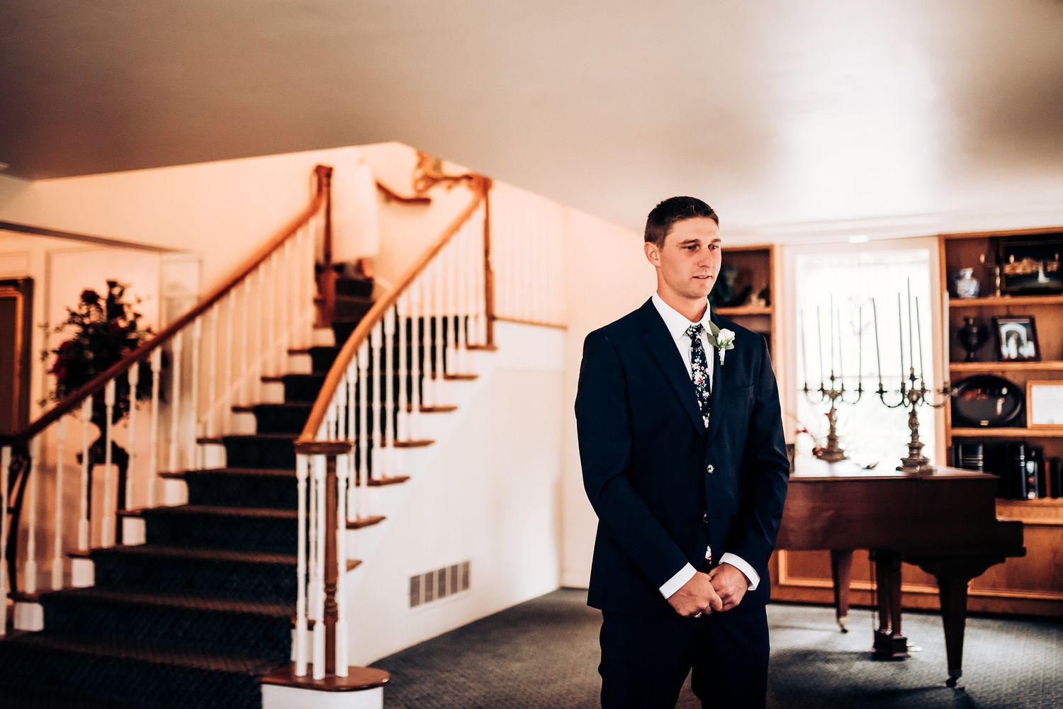 A groom in a classic navy suit awaits within an elegant foyer with a wooden staircase.