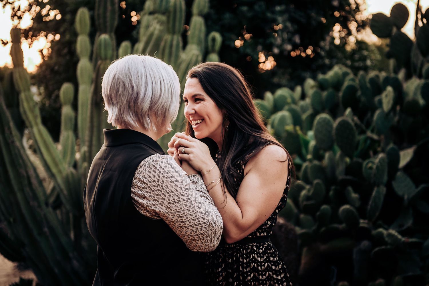 Two people share an intimate laugh while embracing among desert cacti at sunset.
