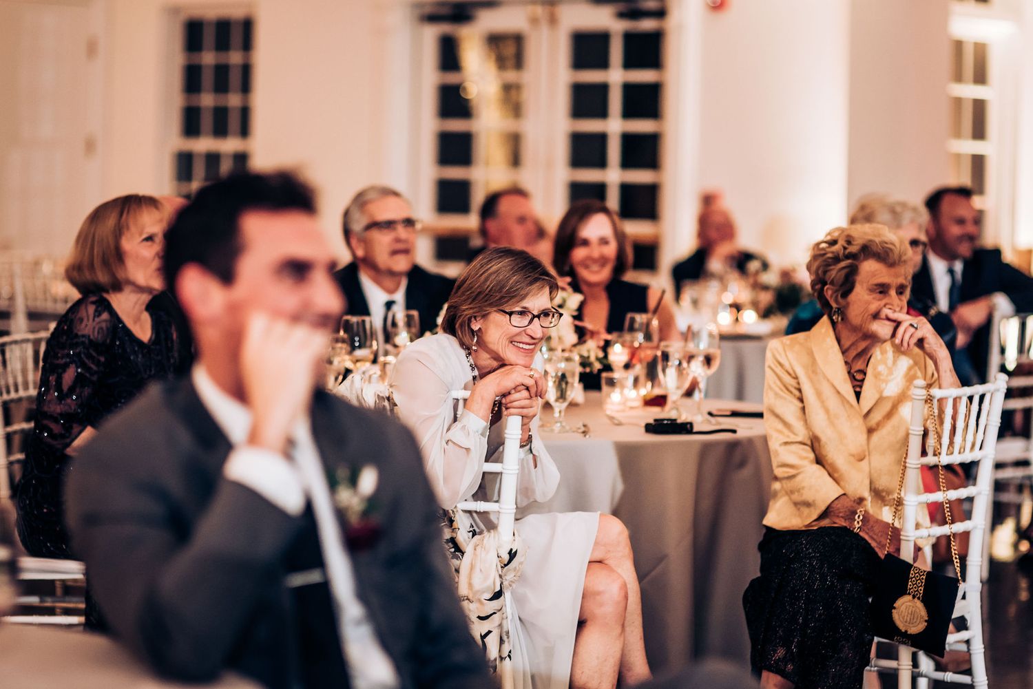 Wedding reception guests seated at round tables listen intently to speeches.