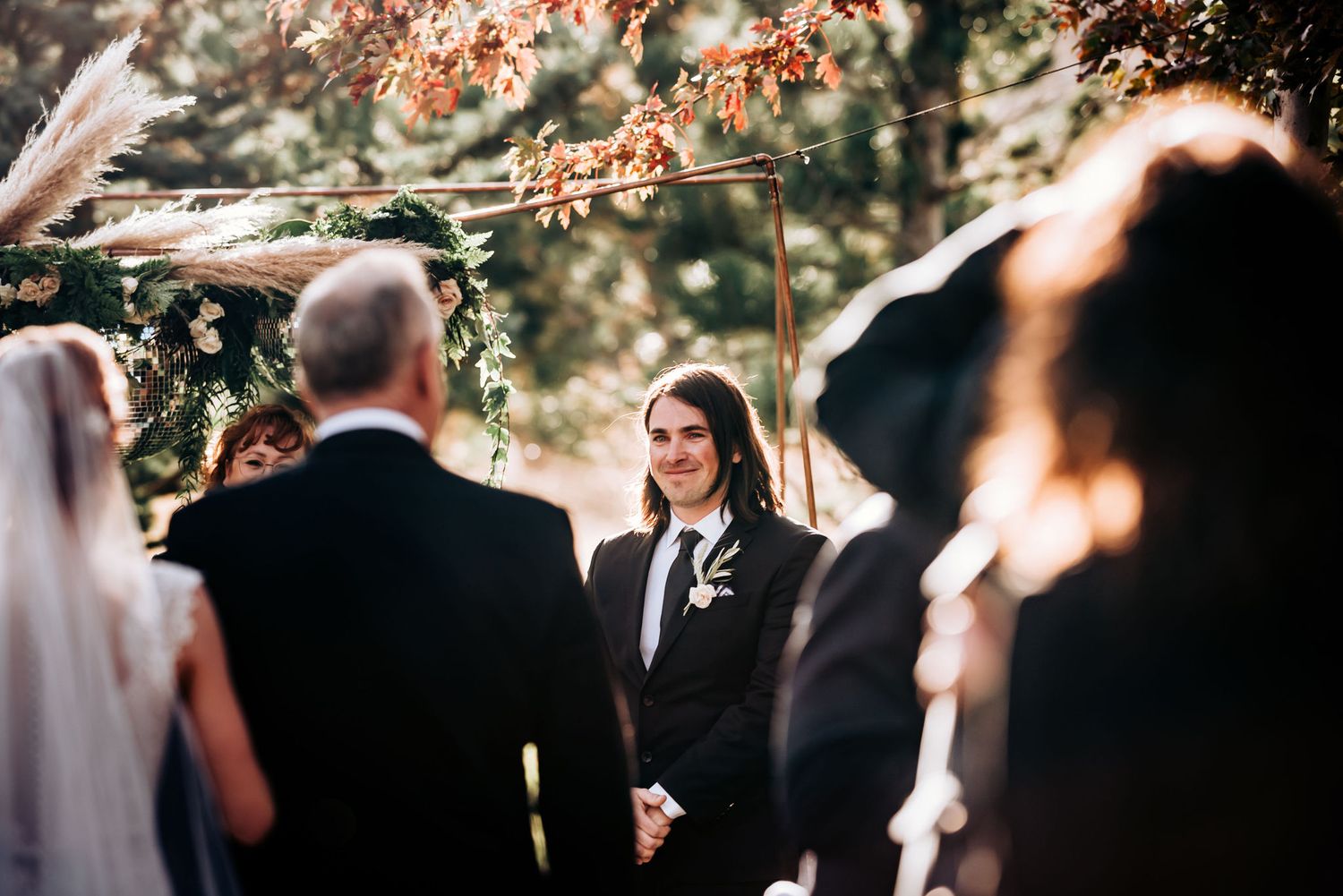 An outdoor wedding ceremony takes place under fall foliage with guests gathered around.