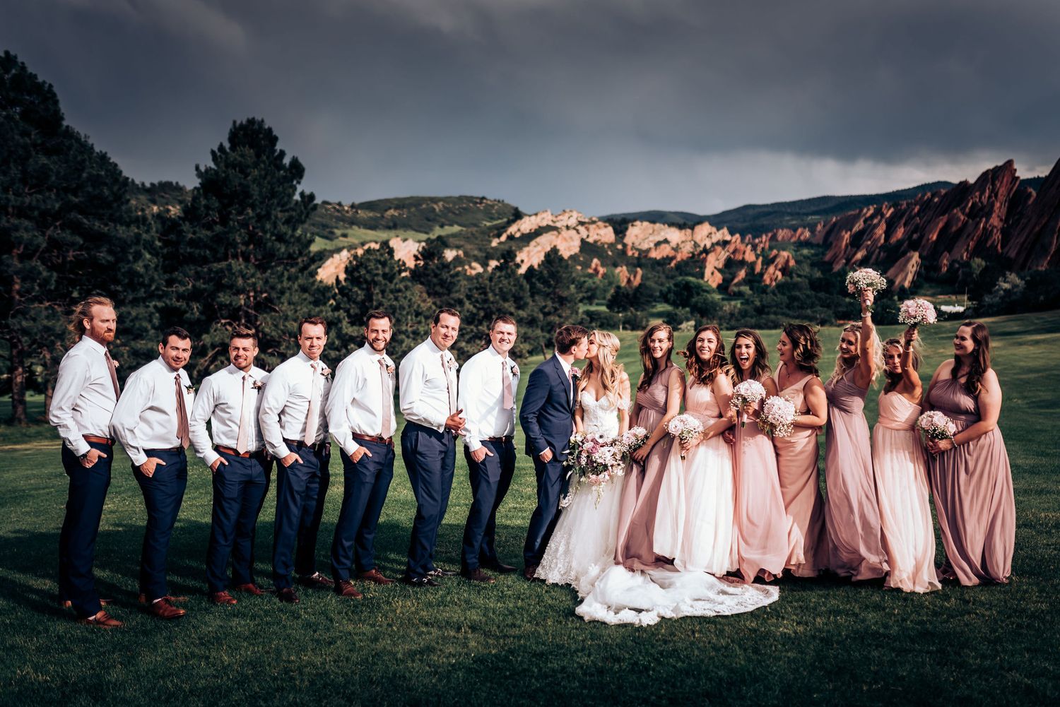Wedding party poses on lawn with dramatic red rock formations and moody sky at Colorado venue.