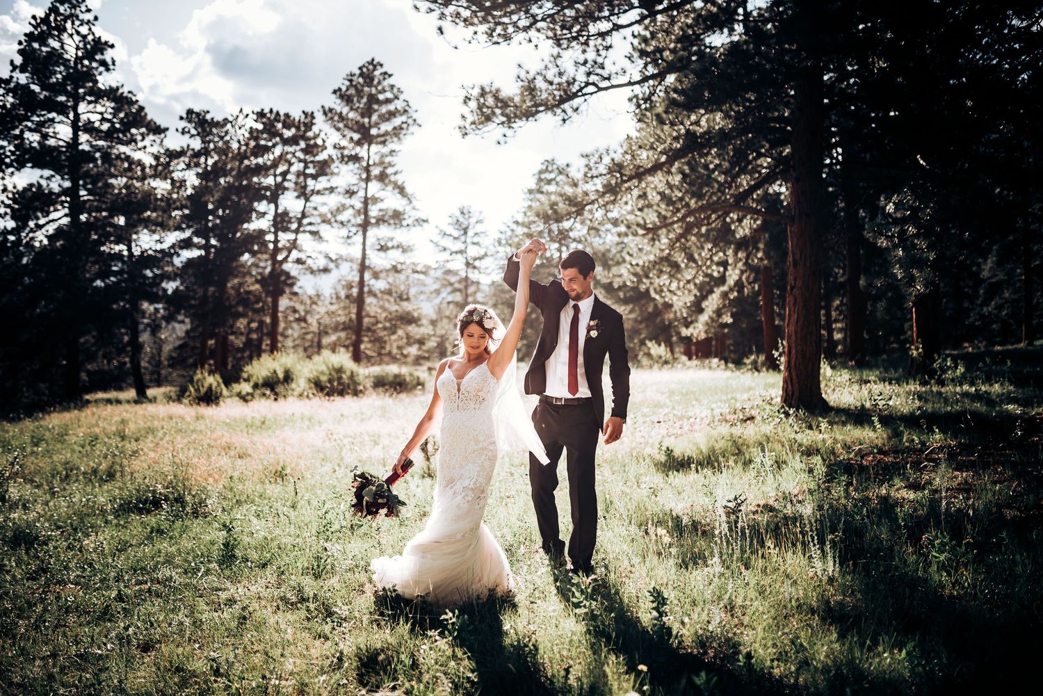 Wedding couple walks hand in hand through sunlit mountain meadow surrounded by tall pine trees.
