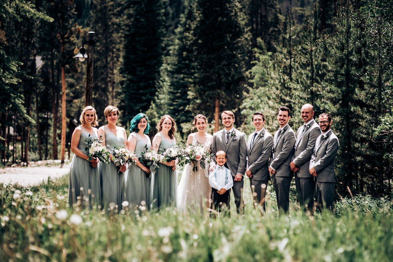 A wedding party in sage green dresses and grey suits poses together in a mountain meadow.