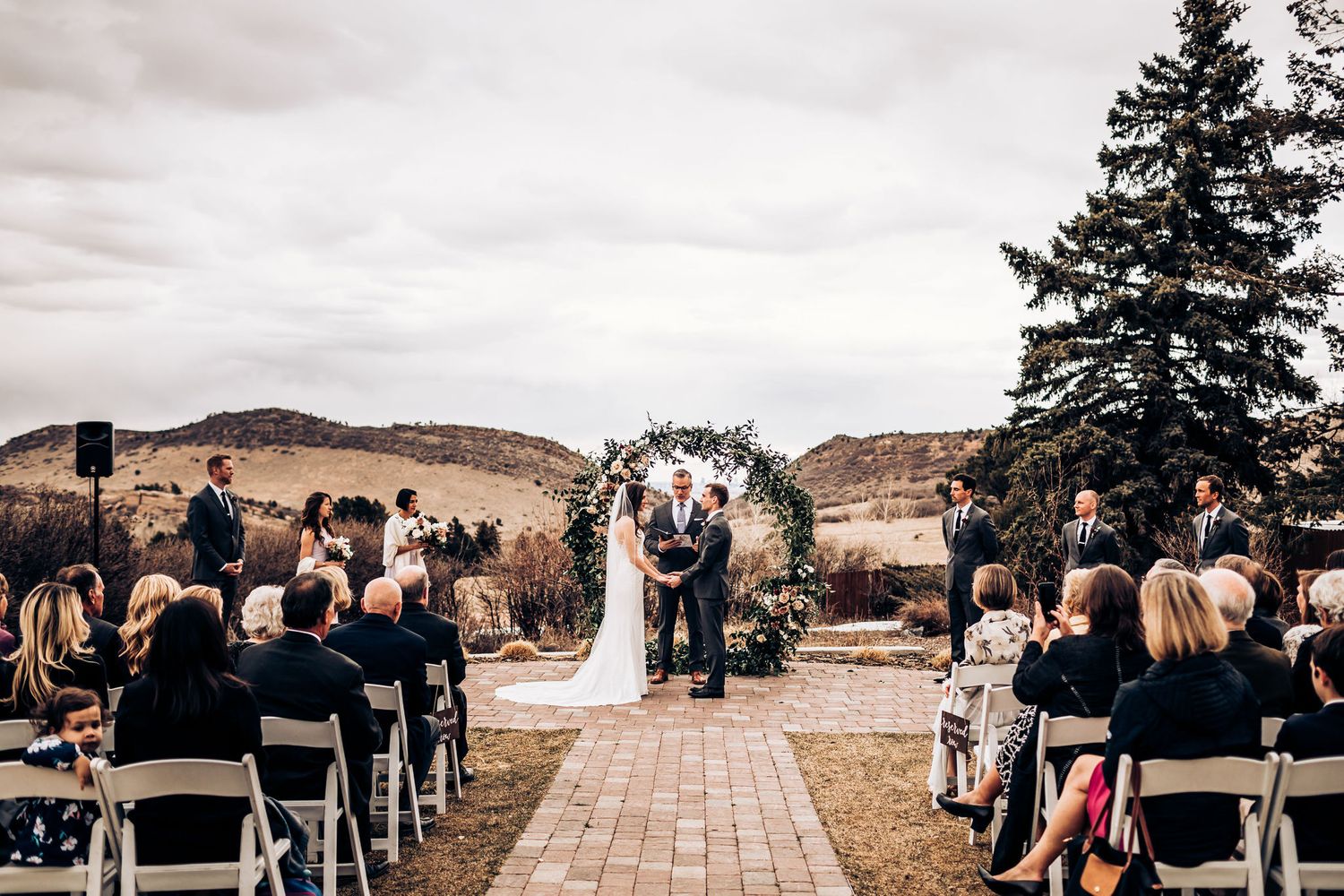 An outdoor wedding ceremony with guests seated on white chairs facing a floral arch against a mountainous backdrop.