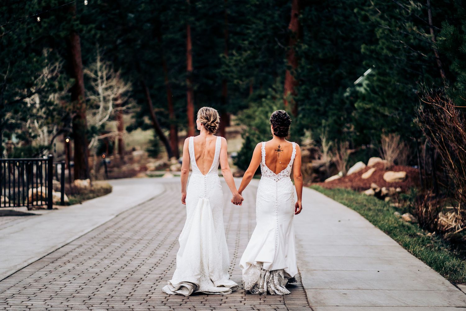Two people in white wedding dresses walking hand in hand on stone path surrounded by trees.