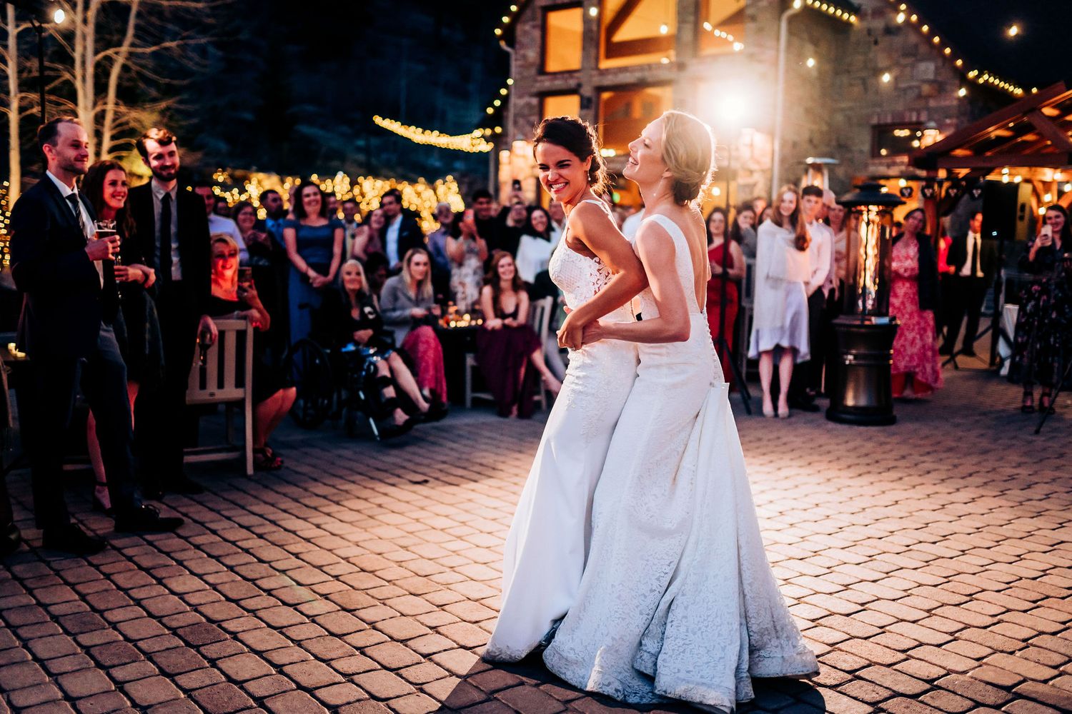 Two people in white dresses share an intimate first dance at an outdoor evening wedding reception.