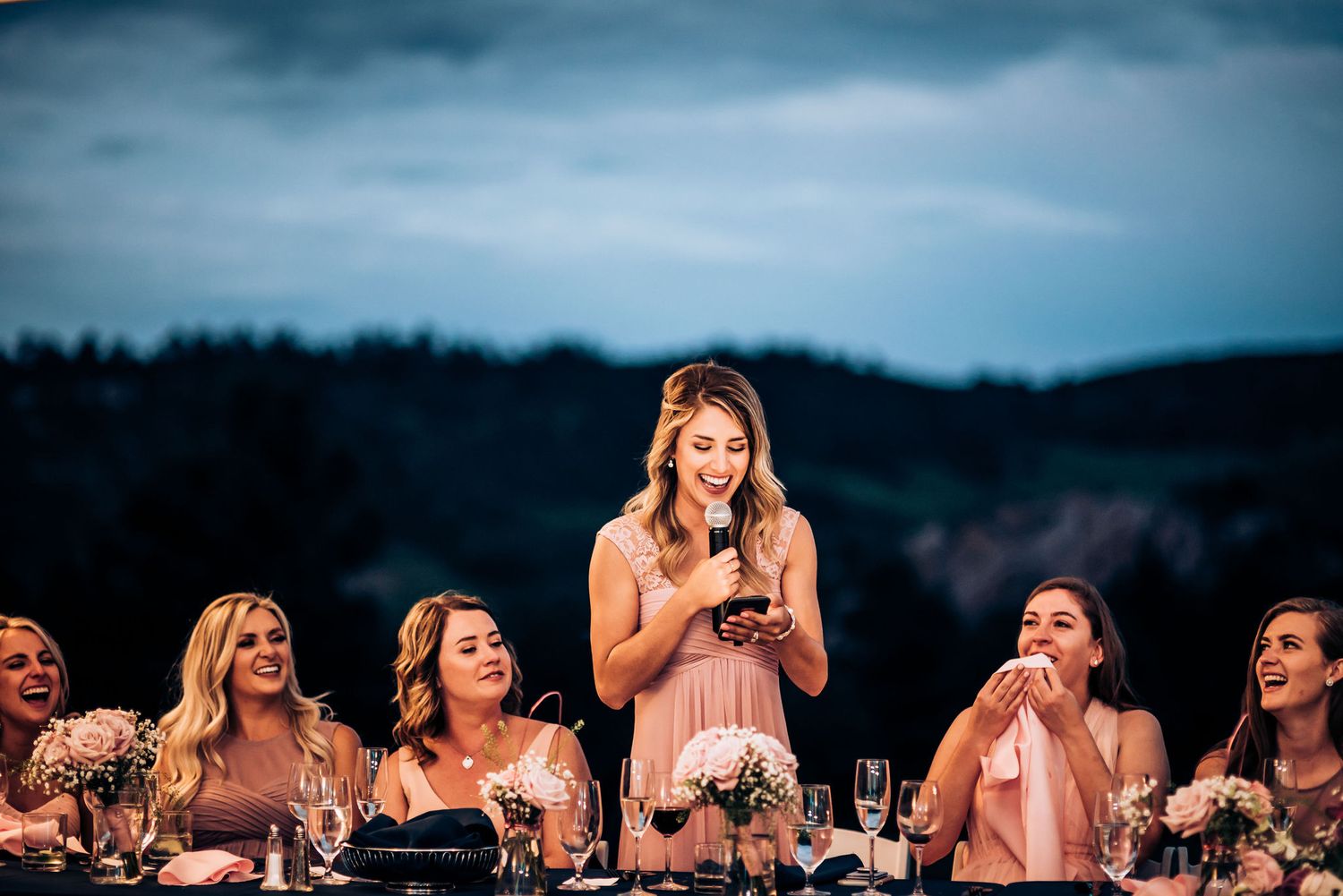 Wedding reception speech moment at outdoor dining table during twilight.