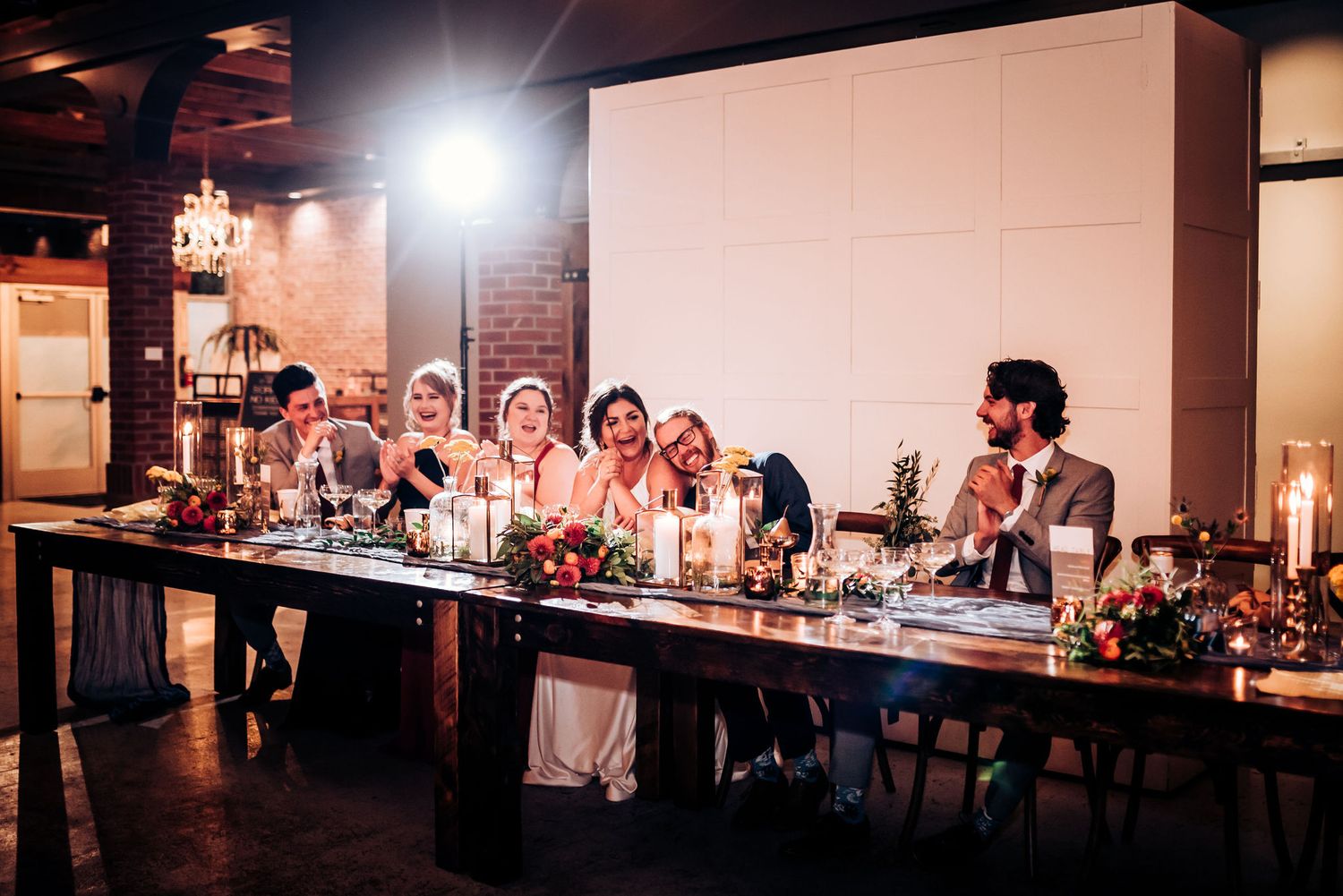 Wedding party shares laughs and conversation at an elegant reception dinner table with rustic brick backdrop.