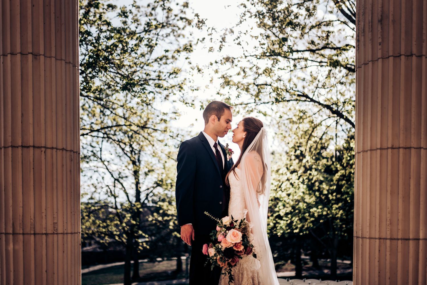 Newlyweds pose between classical columns with sunlight filtering through trees.