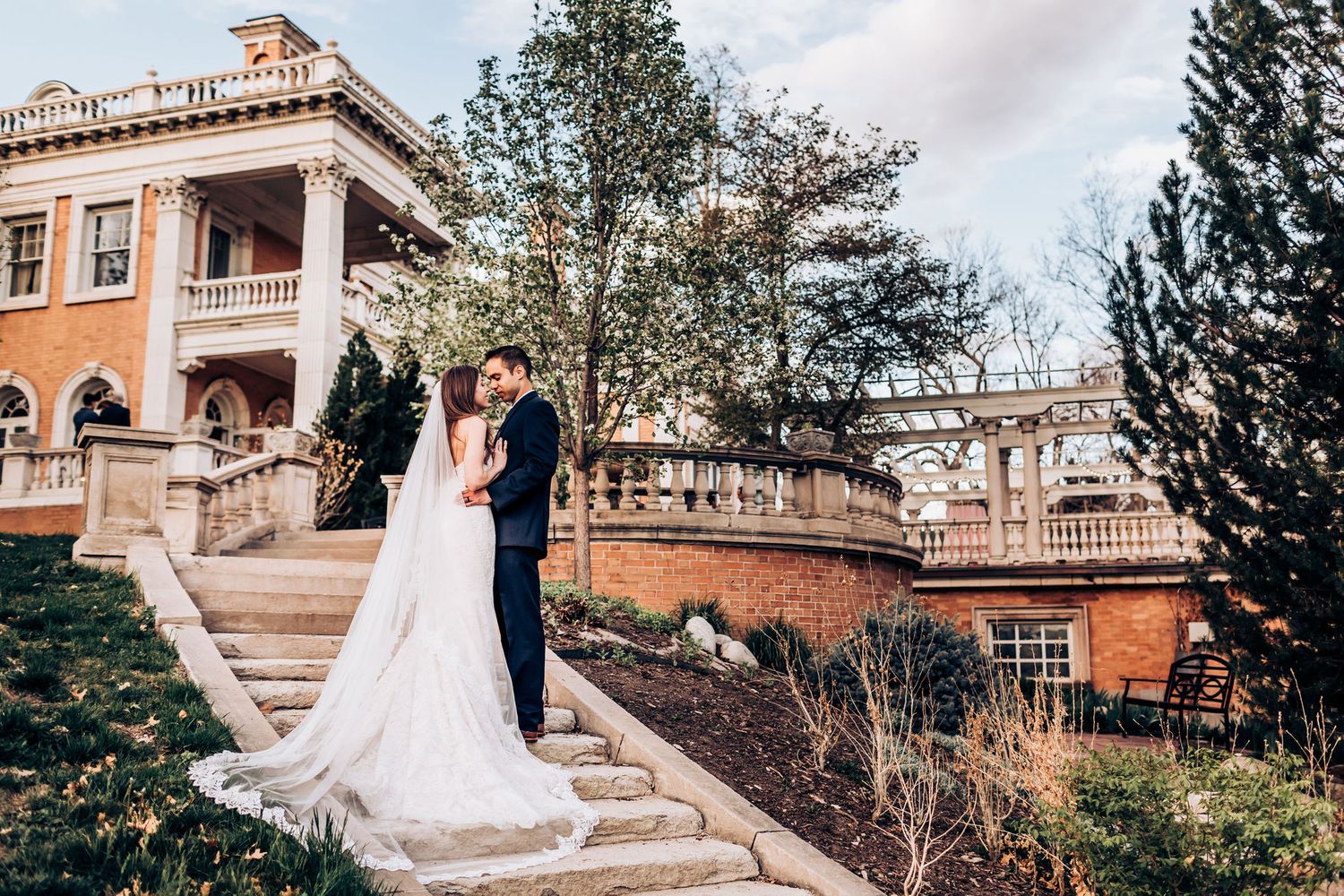 Elegant wedding portrait on mansion staircase with long flowing veil and terracotta architecture.